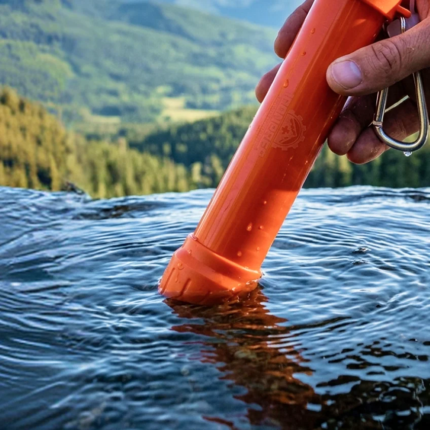 A hand holds an orange portable water filter, dipping it into a stream with mountains in the background.