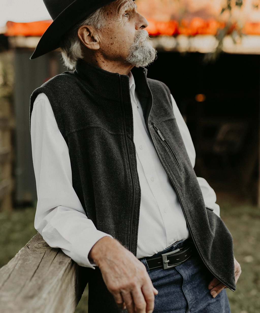 Elderly man in a hat and vest, leaning on a wooden fence.