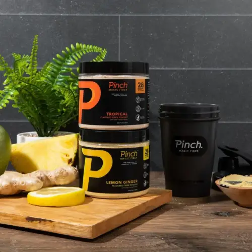 Two jars of Pinch fiber powder and a shaker cup next to pineapple, lemon, and ginger.