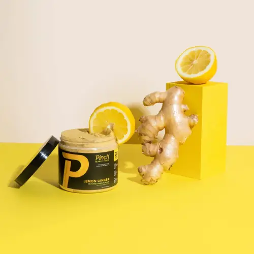 A jar of Pinch Lemon Ginger product with fresh ginger root and lemons on a yellow and white background.