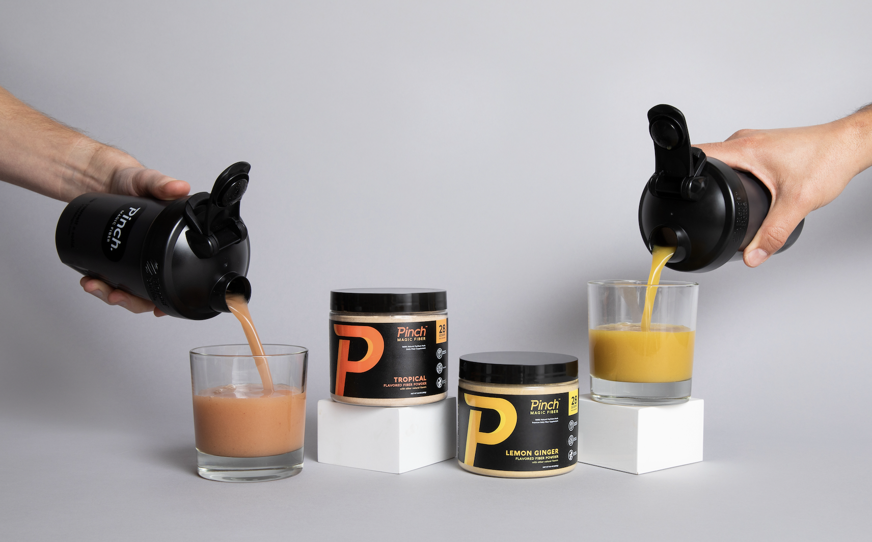 Two hands pouring different flavored Pinch Magic Fiber drinks from shakers into glasses next to the product jars.