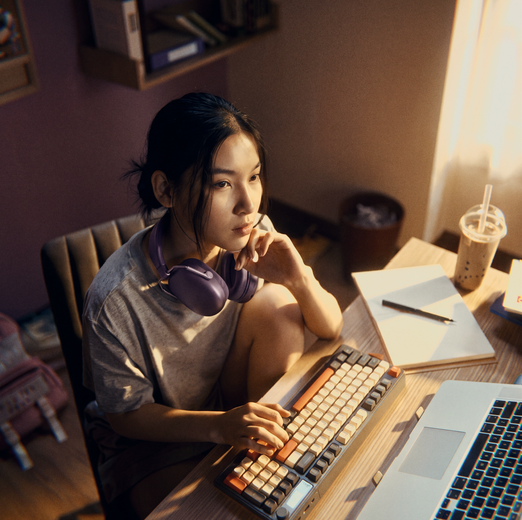 A young woman with headphones around her neck sits thoughtfully at her desk in front of a laptop.