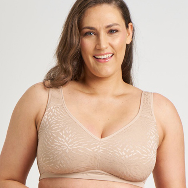 A smiling woman with wavy brown hair models a beige, patterned, soft-cup bra against a light background.