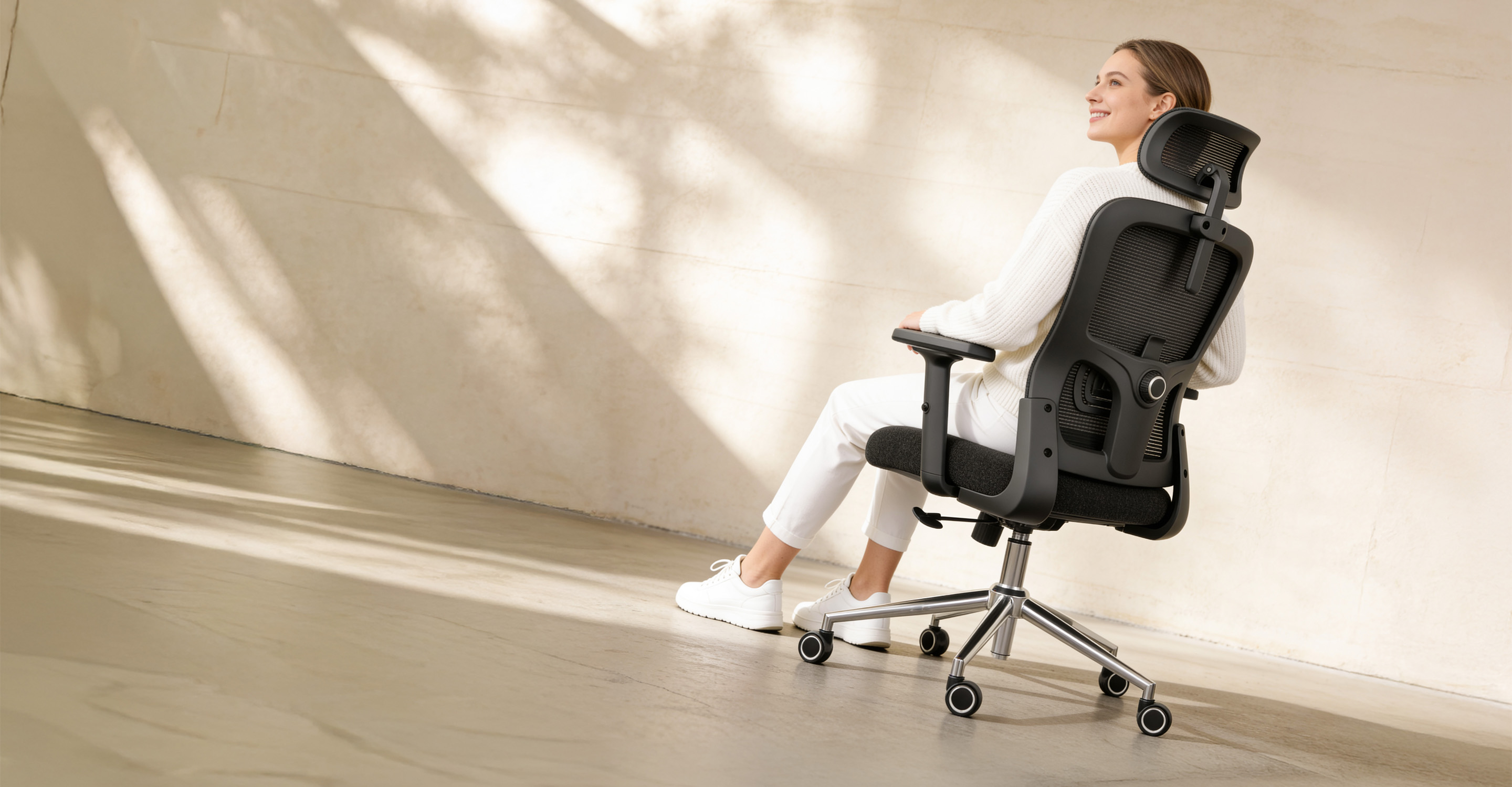 A woman in a white outfit smiles while sitting in a black ergonomic office chair in a sunlit room.