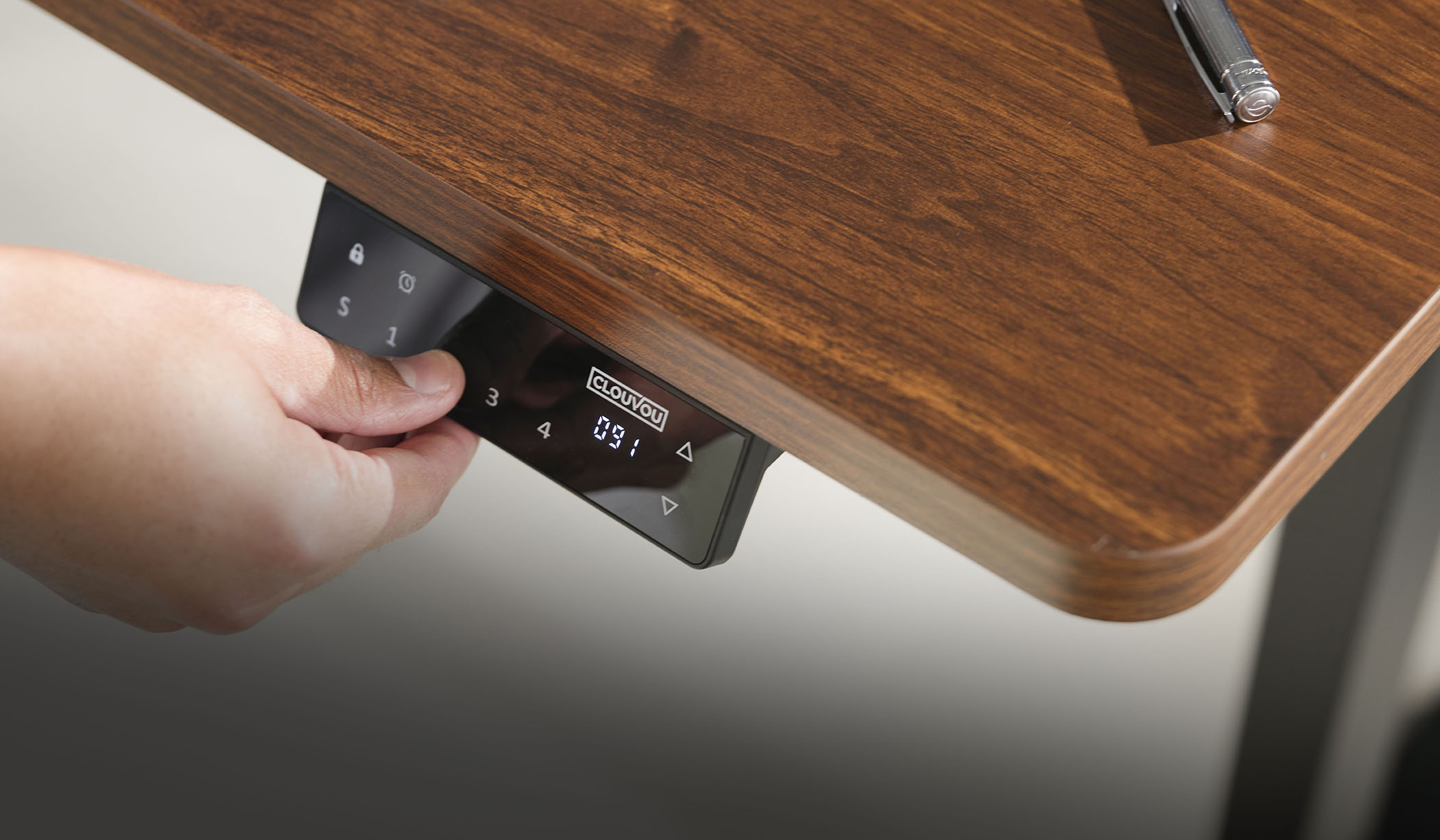 A close-up of a hand adjusting the electronic control panel of a wood-grain standing desk.