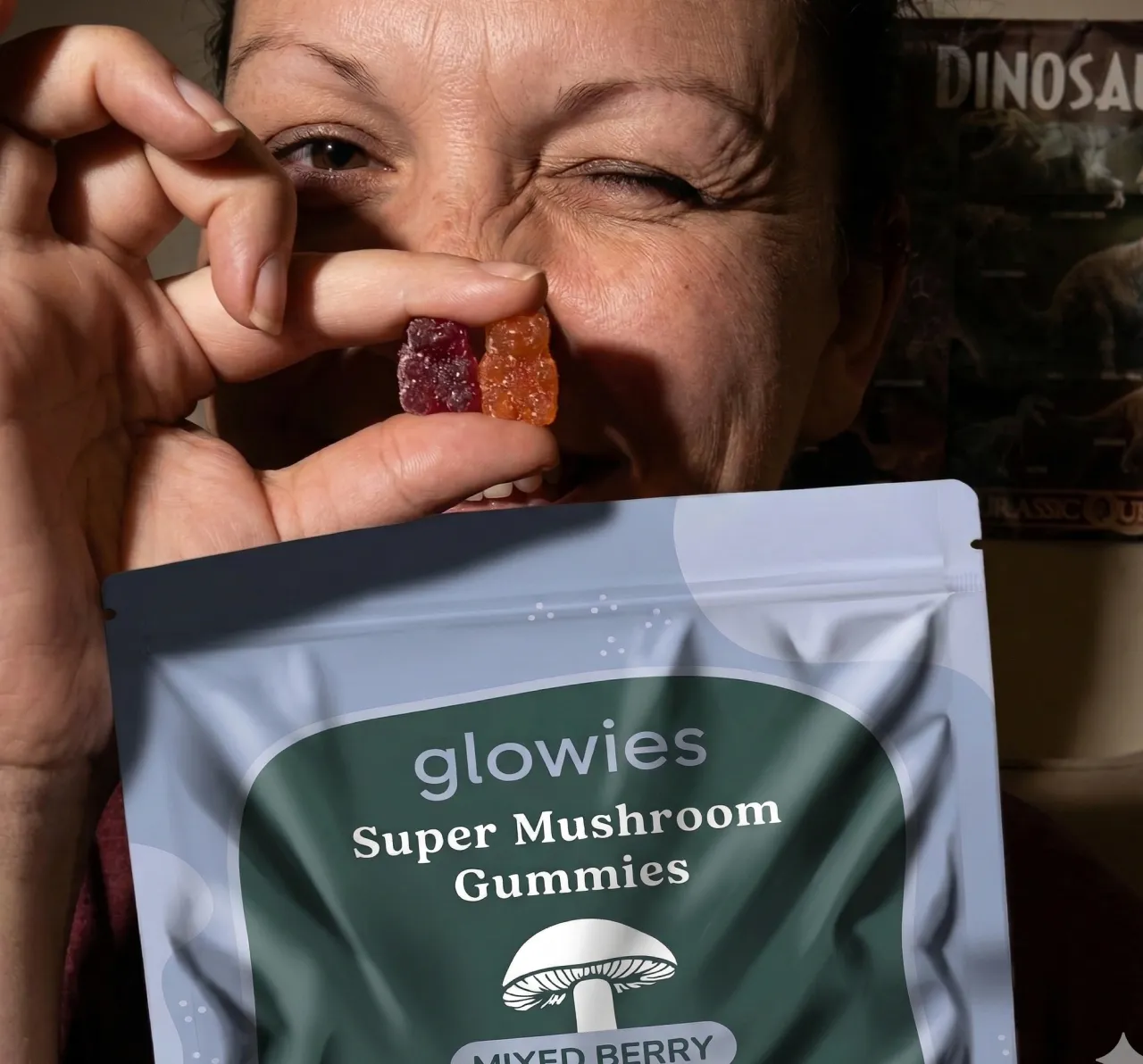 A woman winks while holding two gummy bears and a pouch of Glowies Super Mushroom Gummies.