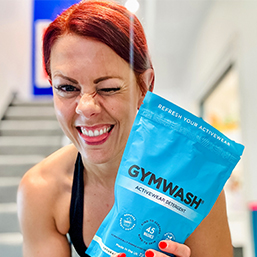 Person winking, holding a blue 'GYMWASH' detergent bag indoors.