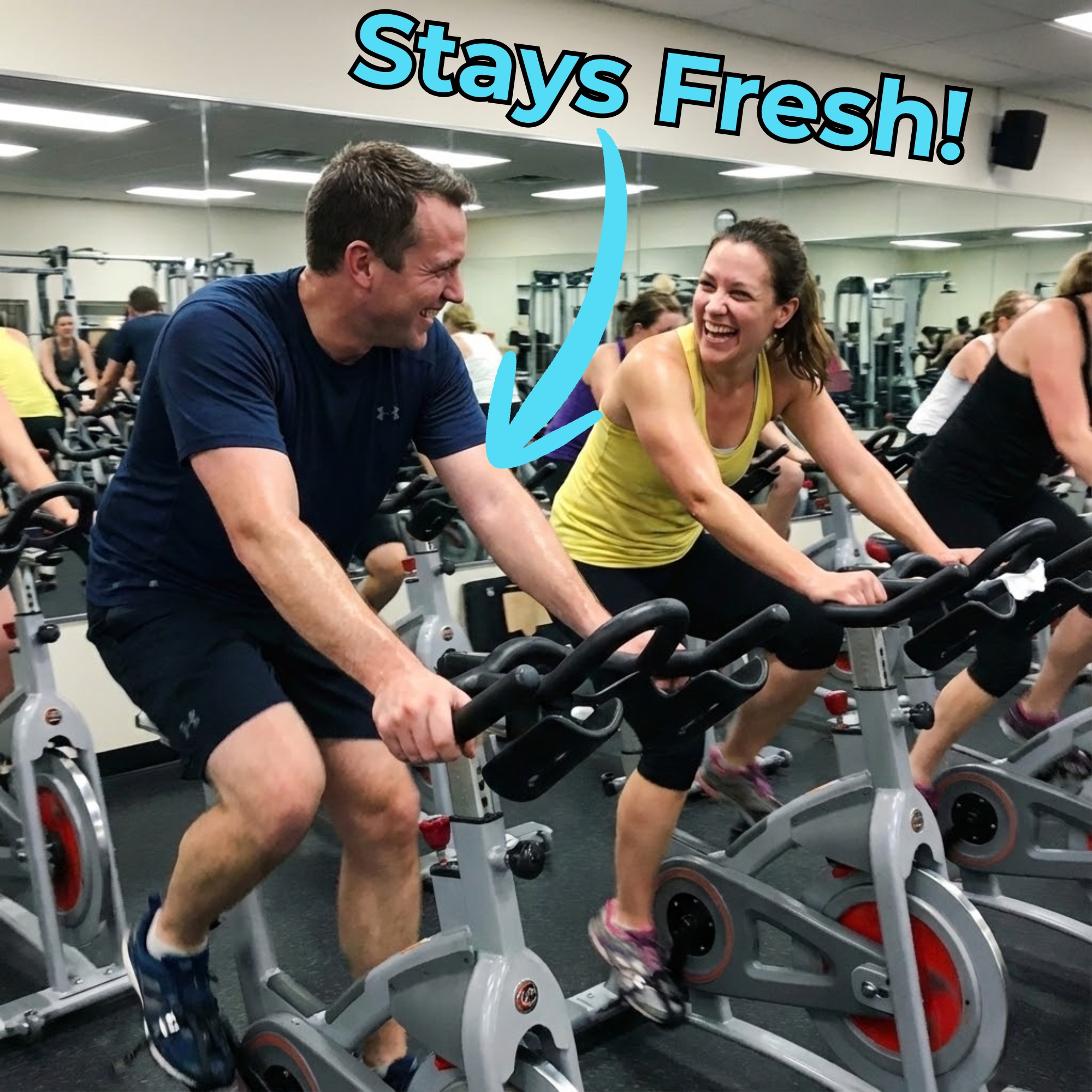 A man and woman laugh together while riding stationary bikes in a crowded gym spin class.