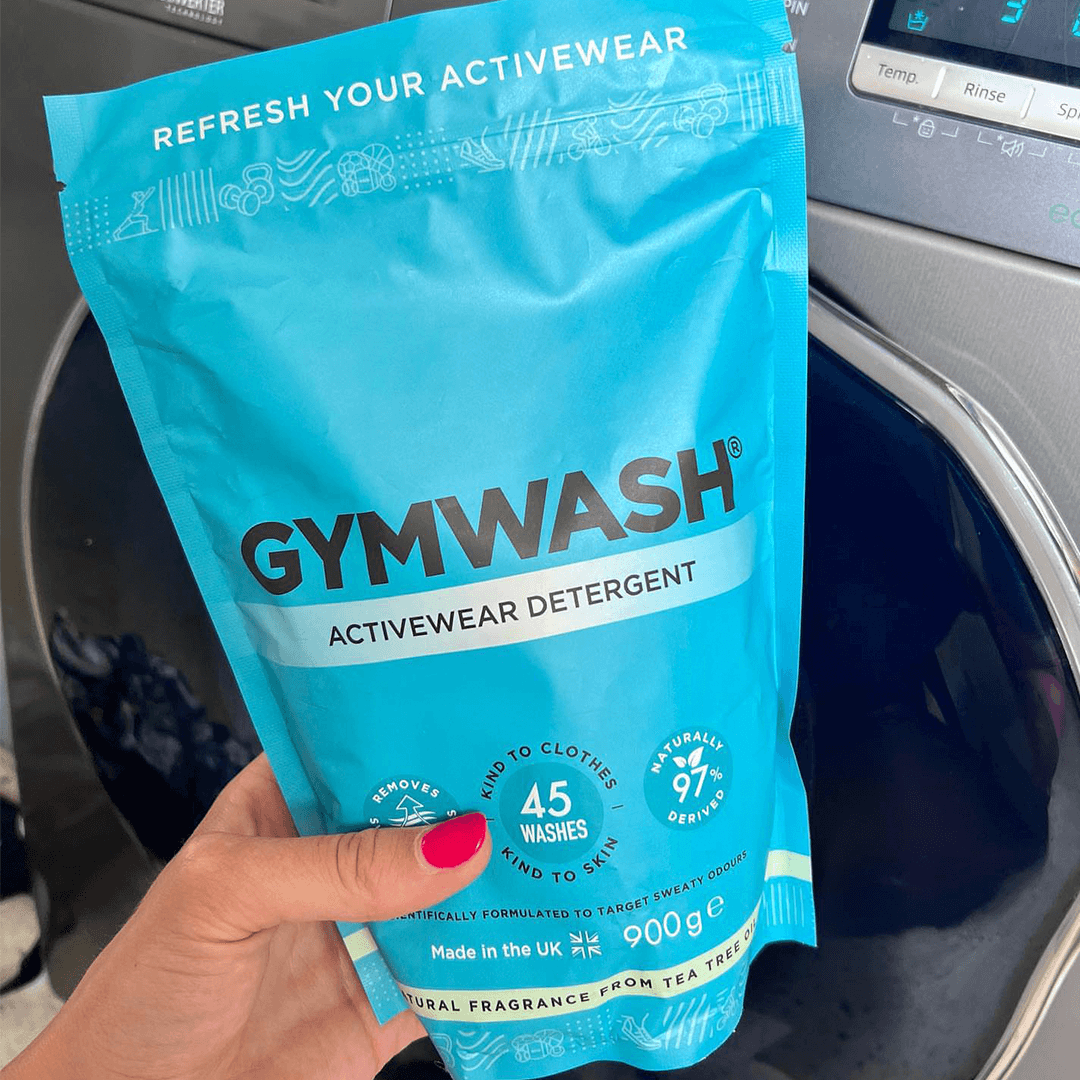 Hand holding a blue Gymwash activewear detergent bag near a washing machine.