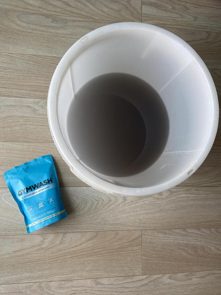 A white bucket with gray water next to a blue detergent packet on wooden floor.