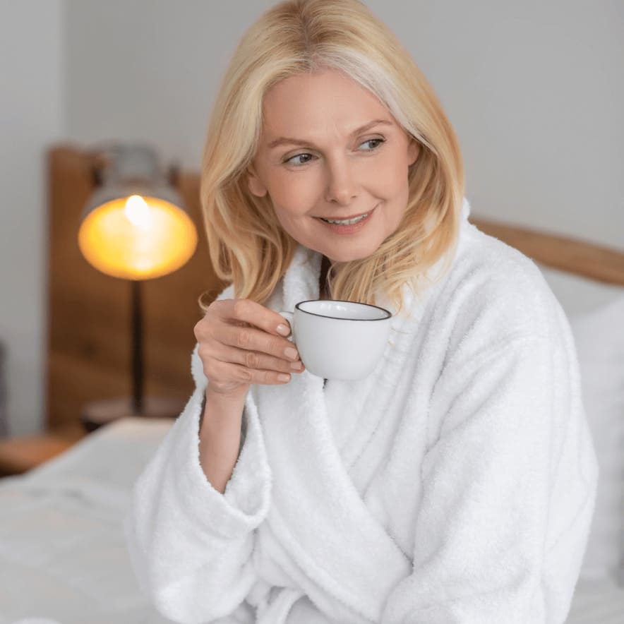 A smiling blonde woman in a white bathrobe sits in a bedroom holding a white cup.