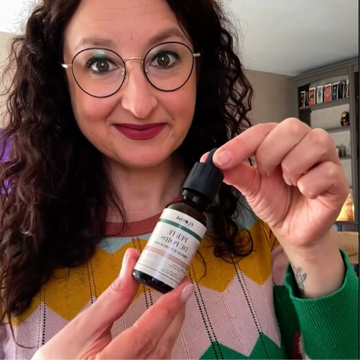 A smiling woman with glasses and dark curly hair holds a small dropper bottle.
