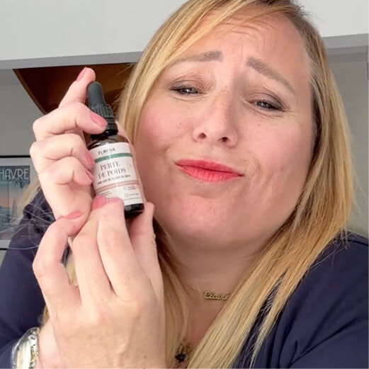 A blonde woman with a skeptical expression holds up a small dropper bottle with a white label.