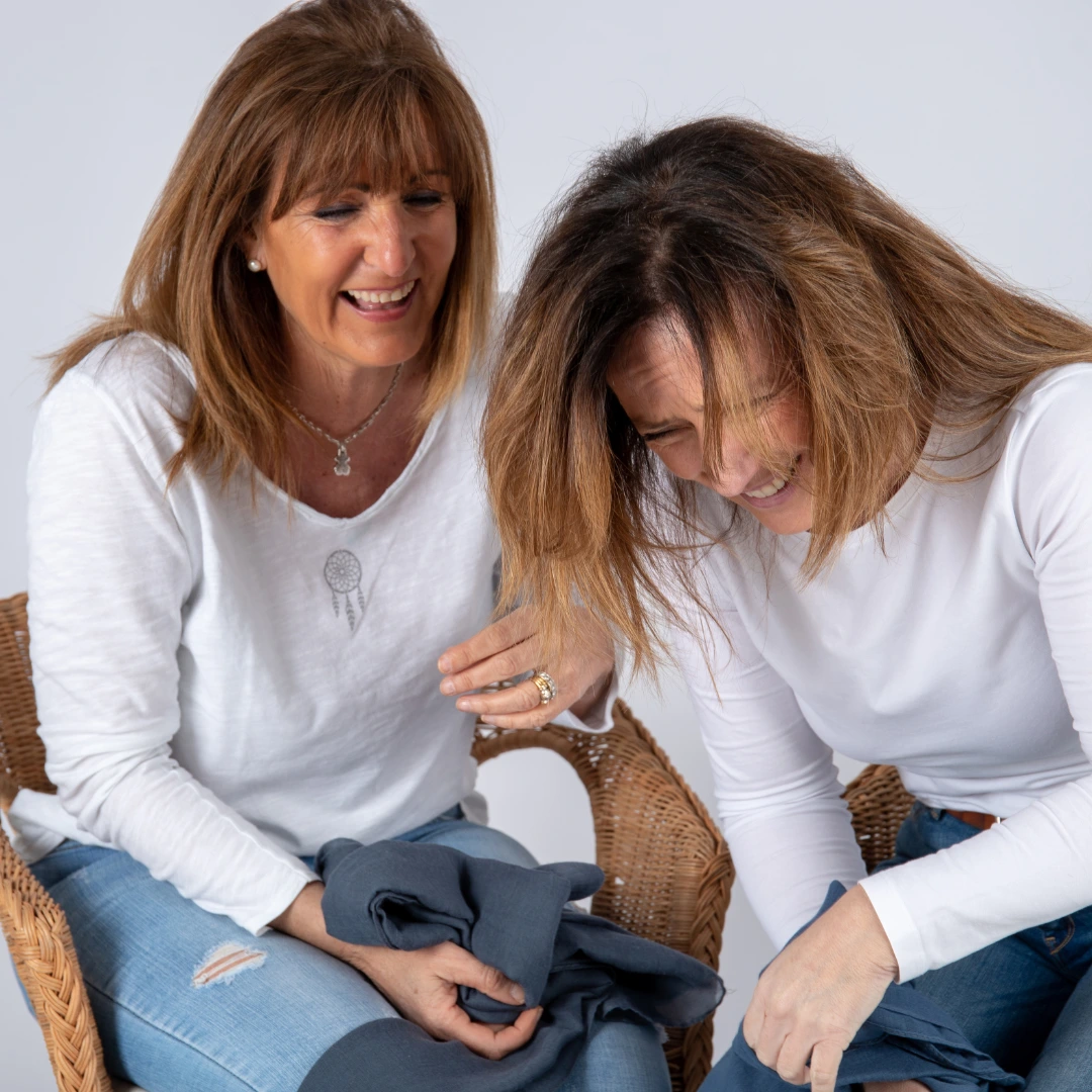 Two women in white shirts and jeans sit in wicker chairs, laughing together against a white background.
