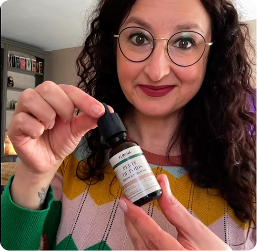 A smiling woman with dark curly hair and glasses holds a small dropper bottle with a white label.