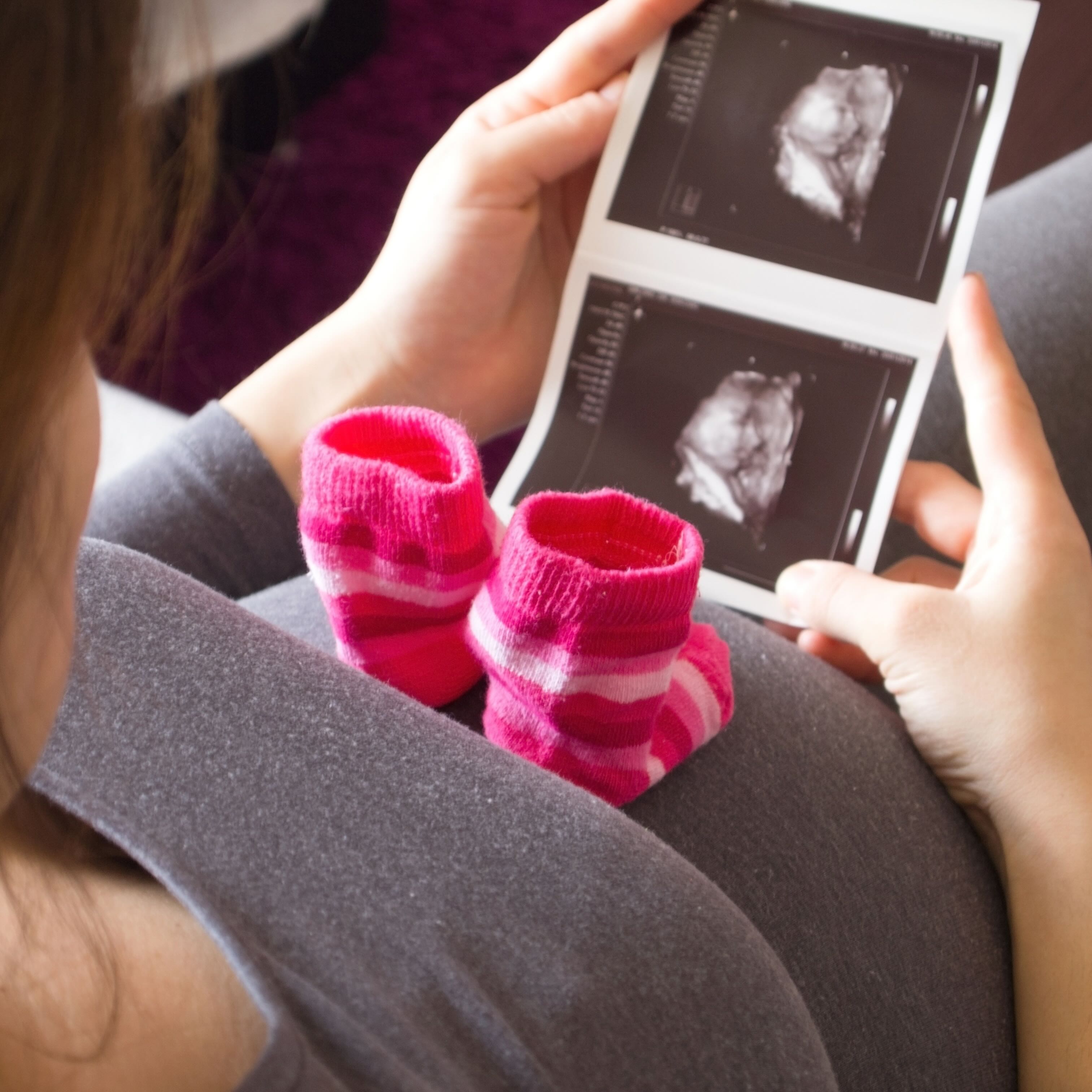 Pregnant woman holding ultrasound images, with pink baby socks on her belly.