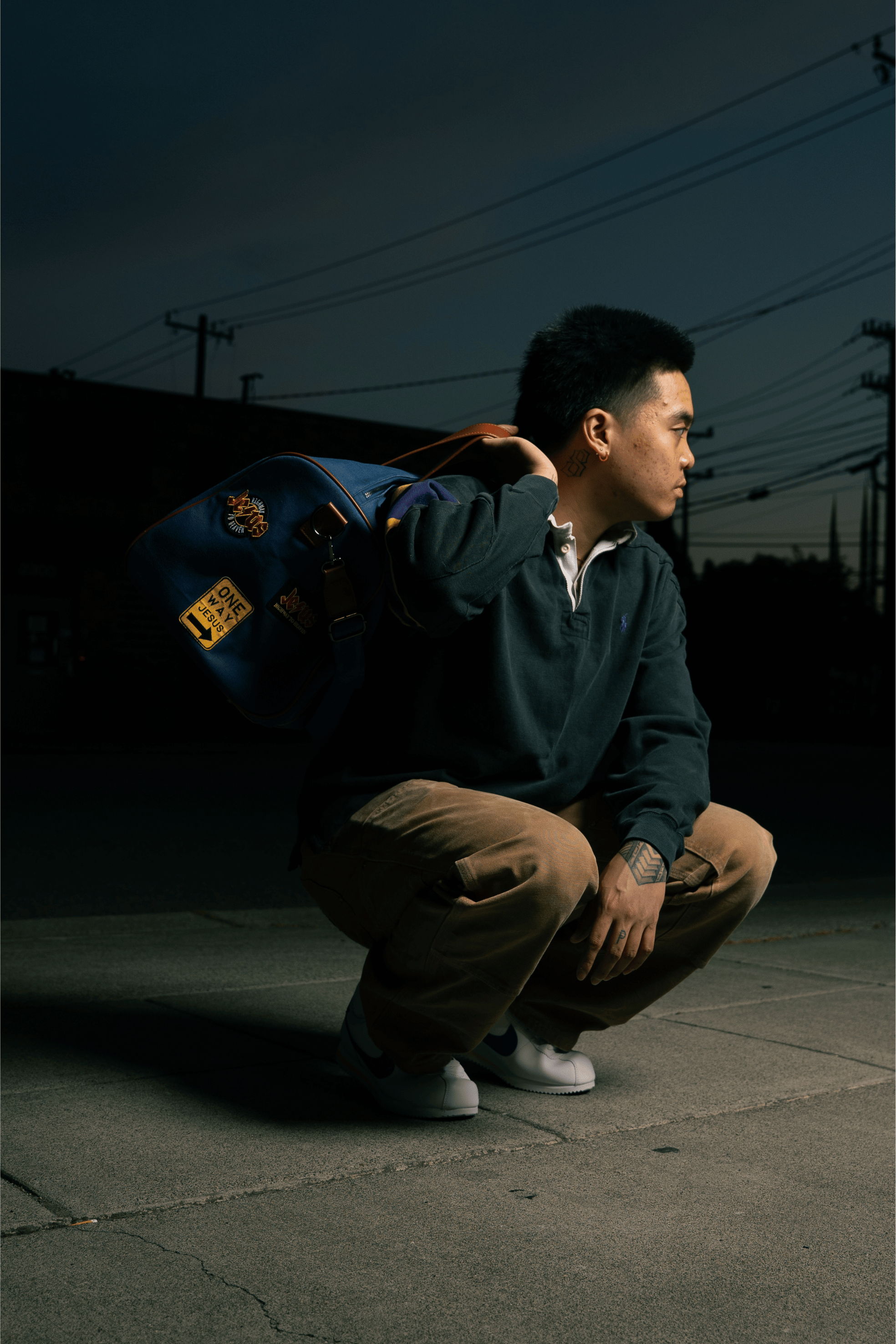Person squatting on sidewalk at dusk, holding a blue bag over shoulder.