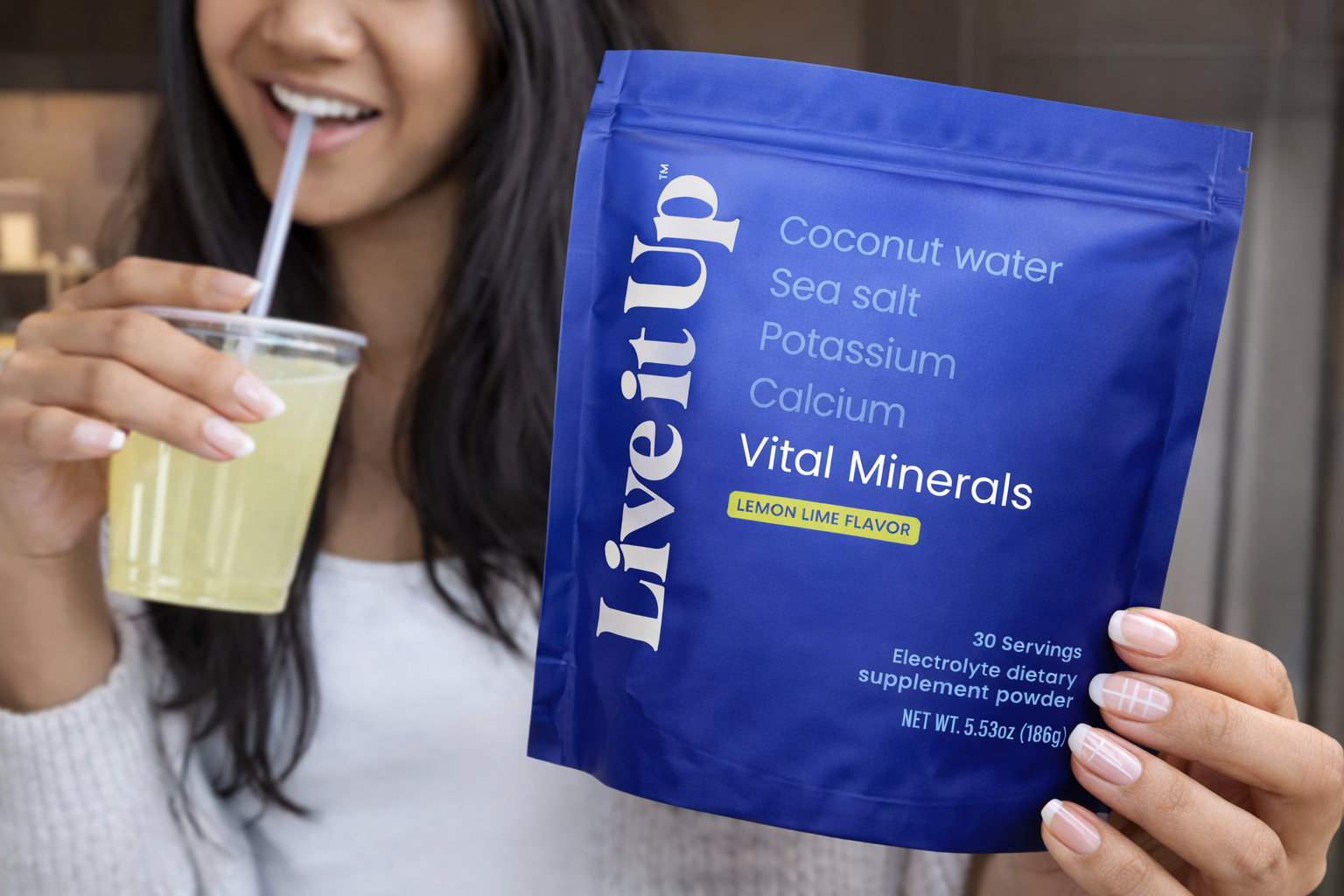 A woman holds a blue pouch of Live it Up Vital Minerals while drinking a yellow beverage.