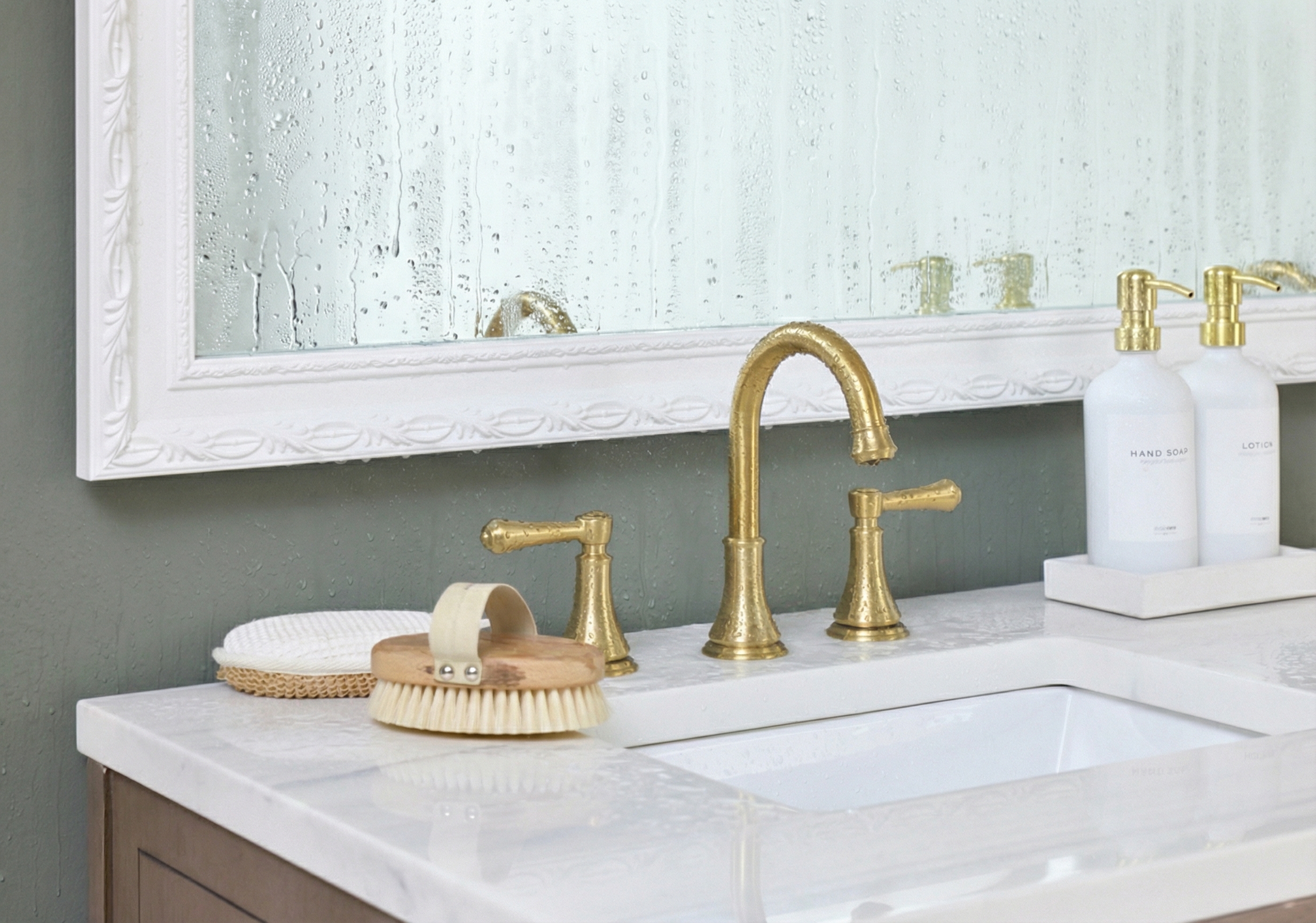 A gold faucet on a marble bathroom vanity, with a mirror and countertop covered in water droplets.