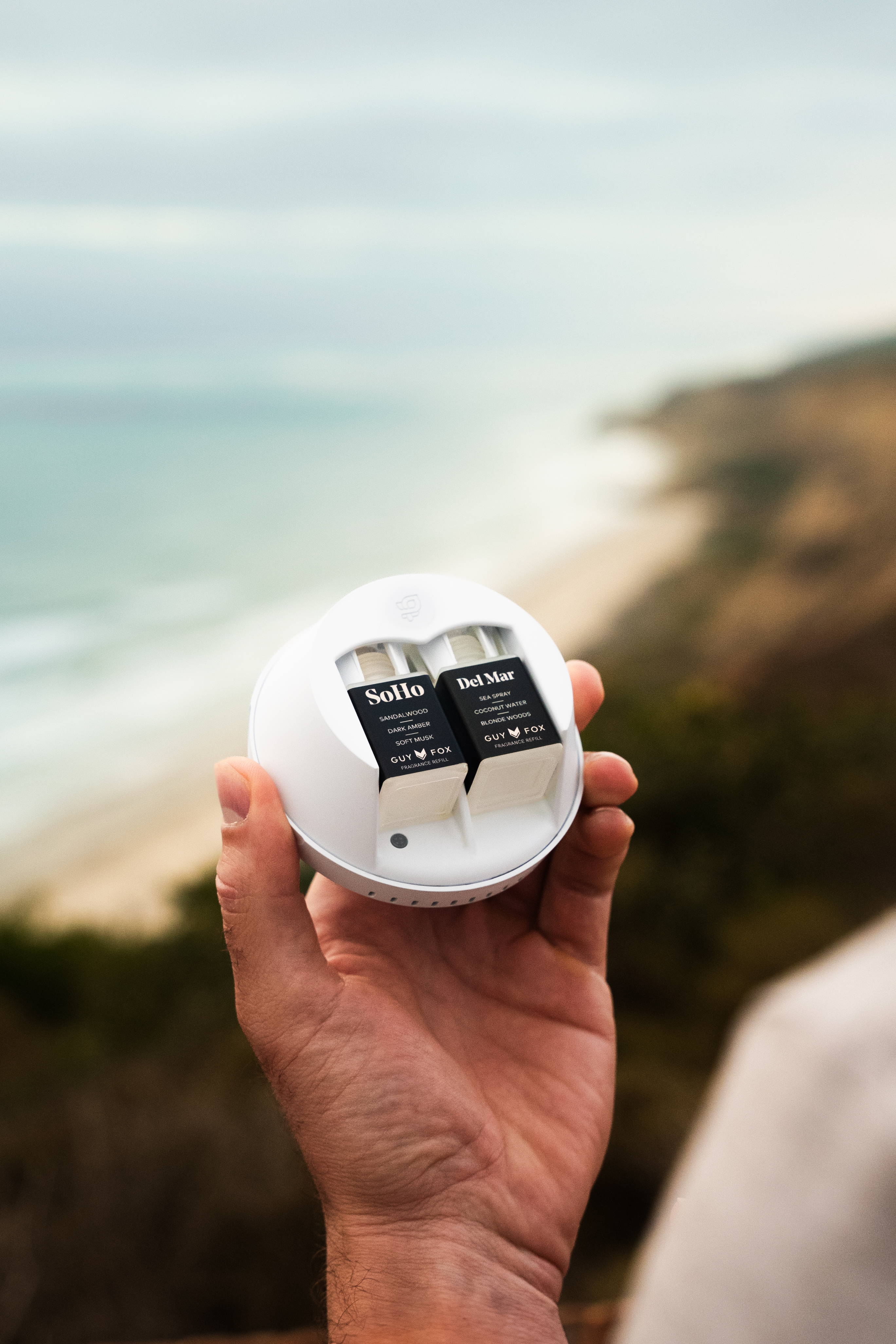 Hand holding a round device with two fragrance cartridges, beach in background.