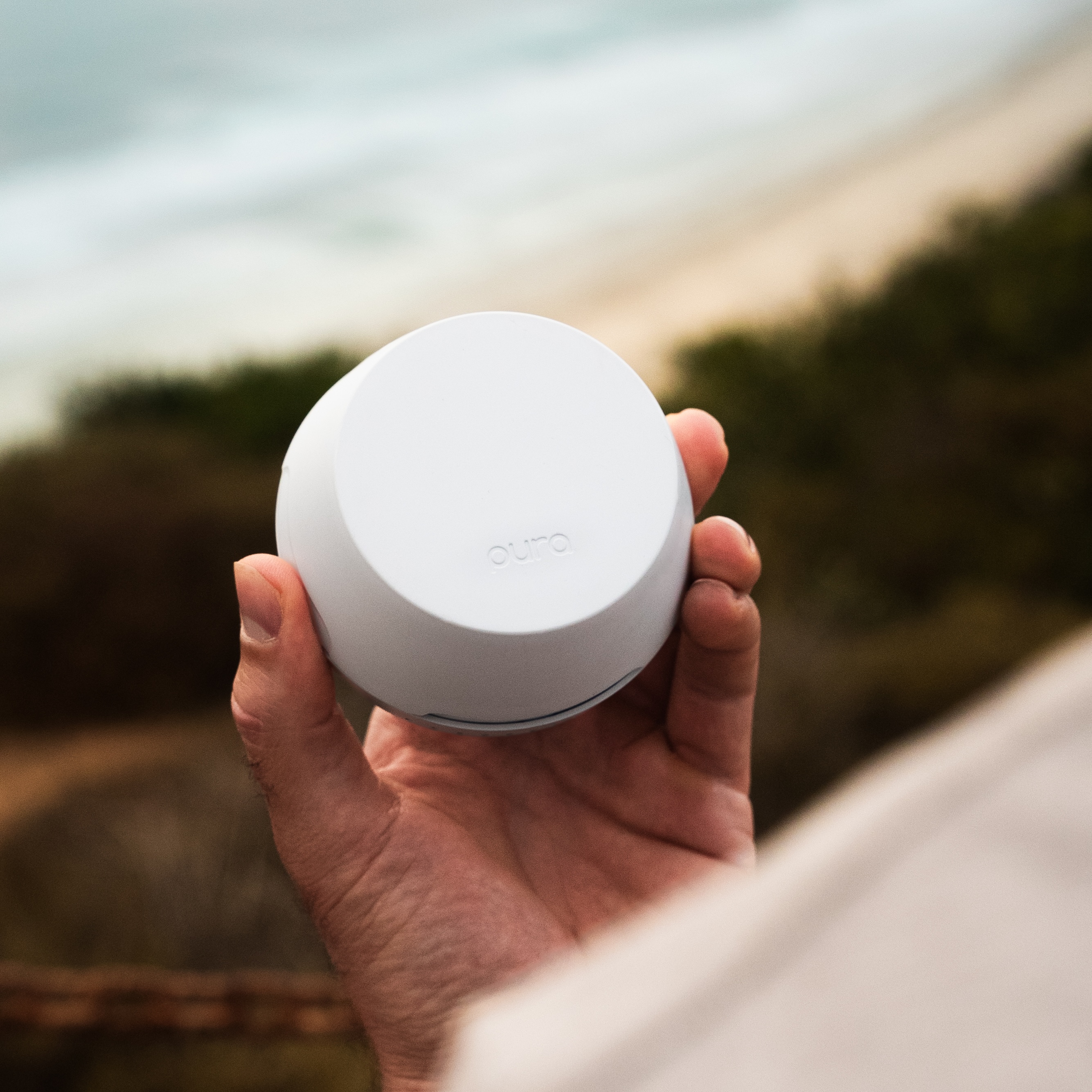 Hand holding a white circular device with 'oura' text, blurred background.