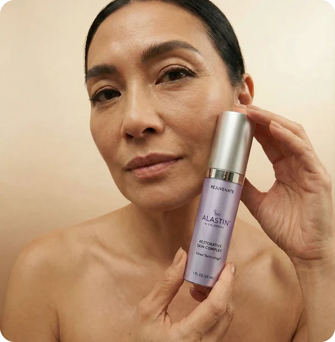 A woman with dark hair holds a bottle of Alastin skincare product next to her face.