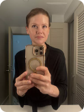 A woman with short, pulled-back hair wearing a black top takes a selfie in a mirror.