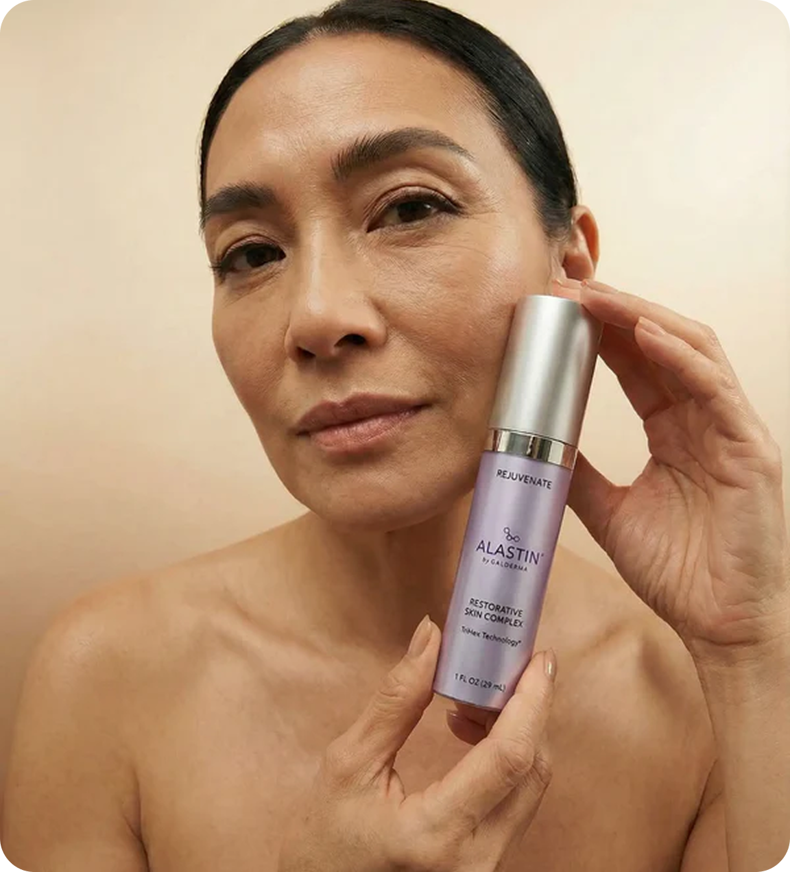 A woman with her hair pulled back holds a bottle of Alastin skincare product to her cheek.