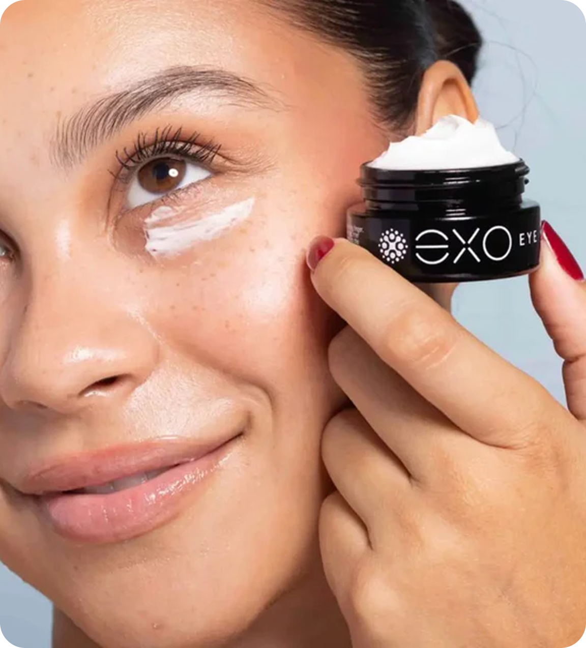 A woman applies white EXO EYE cream under her eye from a small black jar.