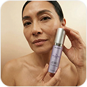 A woman holds a purple and silver bottle of a skincare product next to her face.