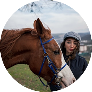 A woman in a grey cap stands next to a brown and white horse with a blue bridle.