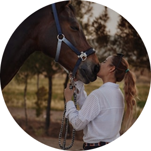 A woman with a long ponytail kisses a brown horse on the nose outdoors.