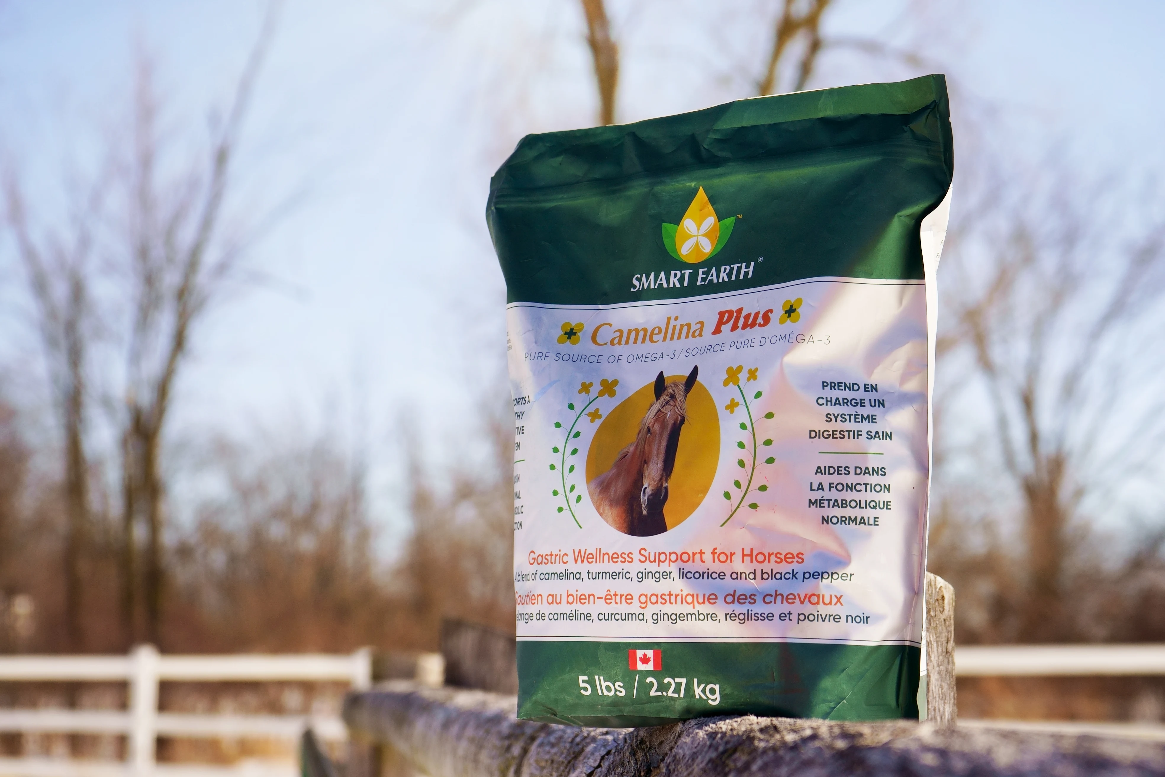 A bag of Smart Earth Camelina Plus horse supplement resting on a wooden fence outdoors.