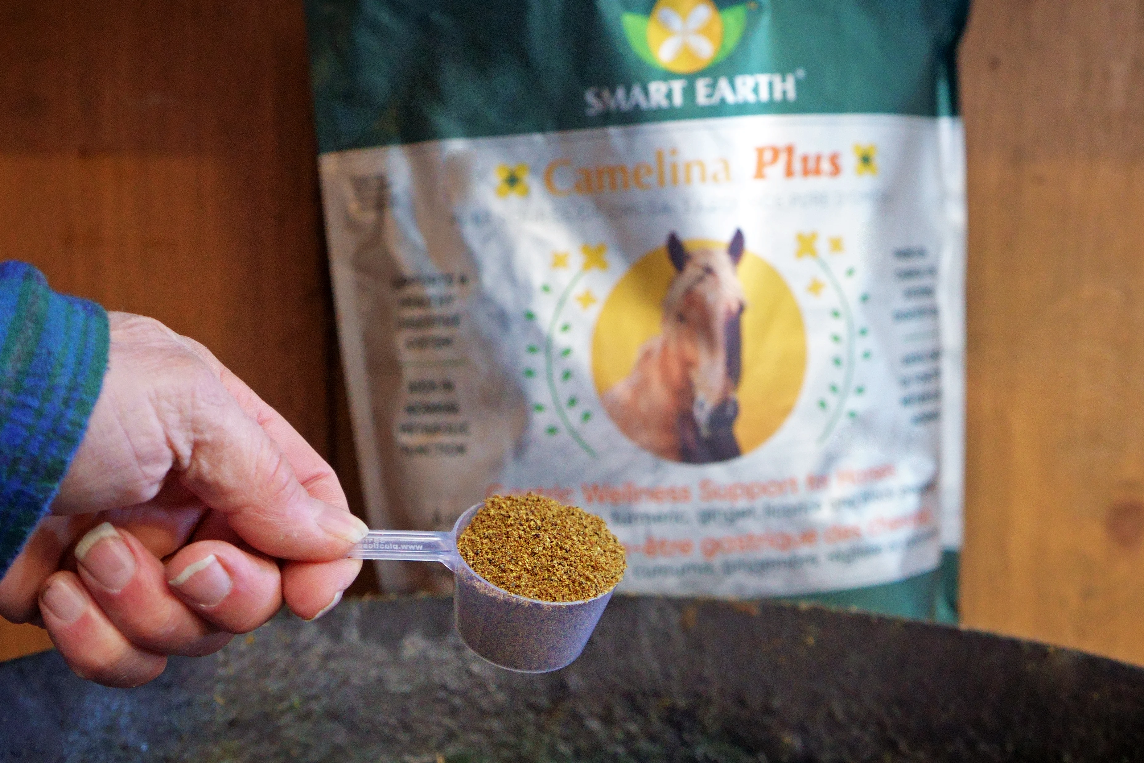 A hand holds a scoop of Smart Earth Camelina Plus horse supplement in front of the bag.