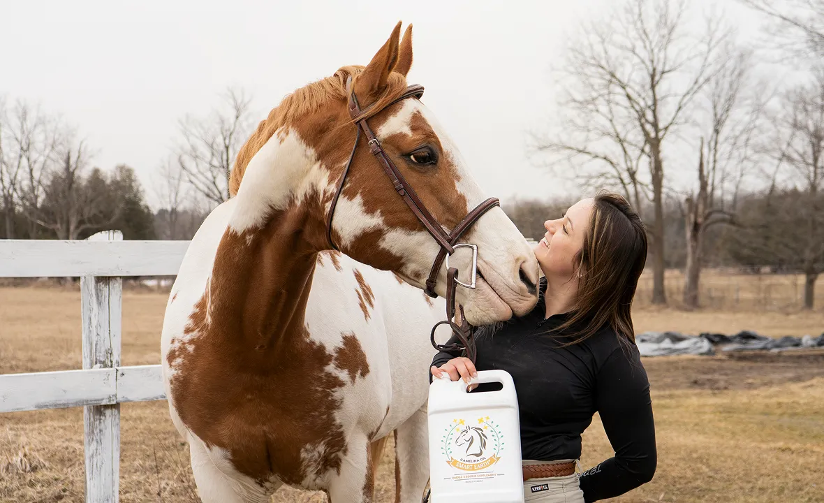 A woman holding a white jug stands with her brown and white horse, nuzzling its face.