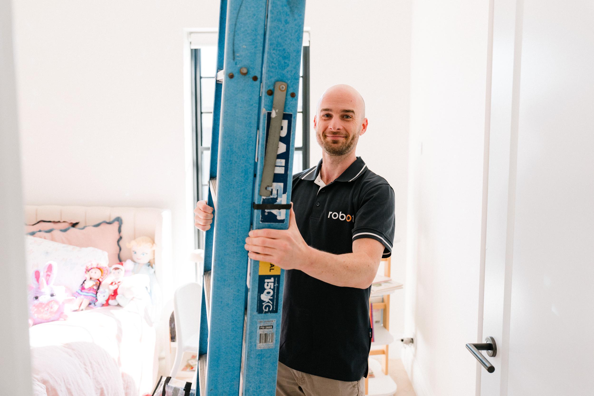 Man holding a blue ladder in a bright room with toys on a bed.