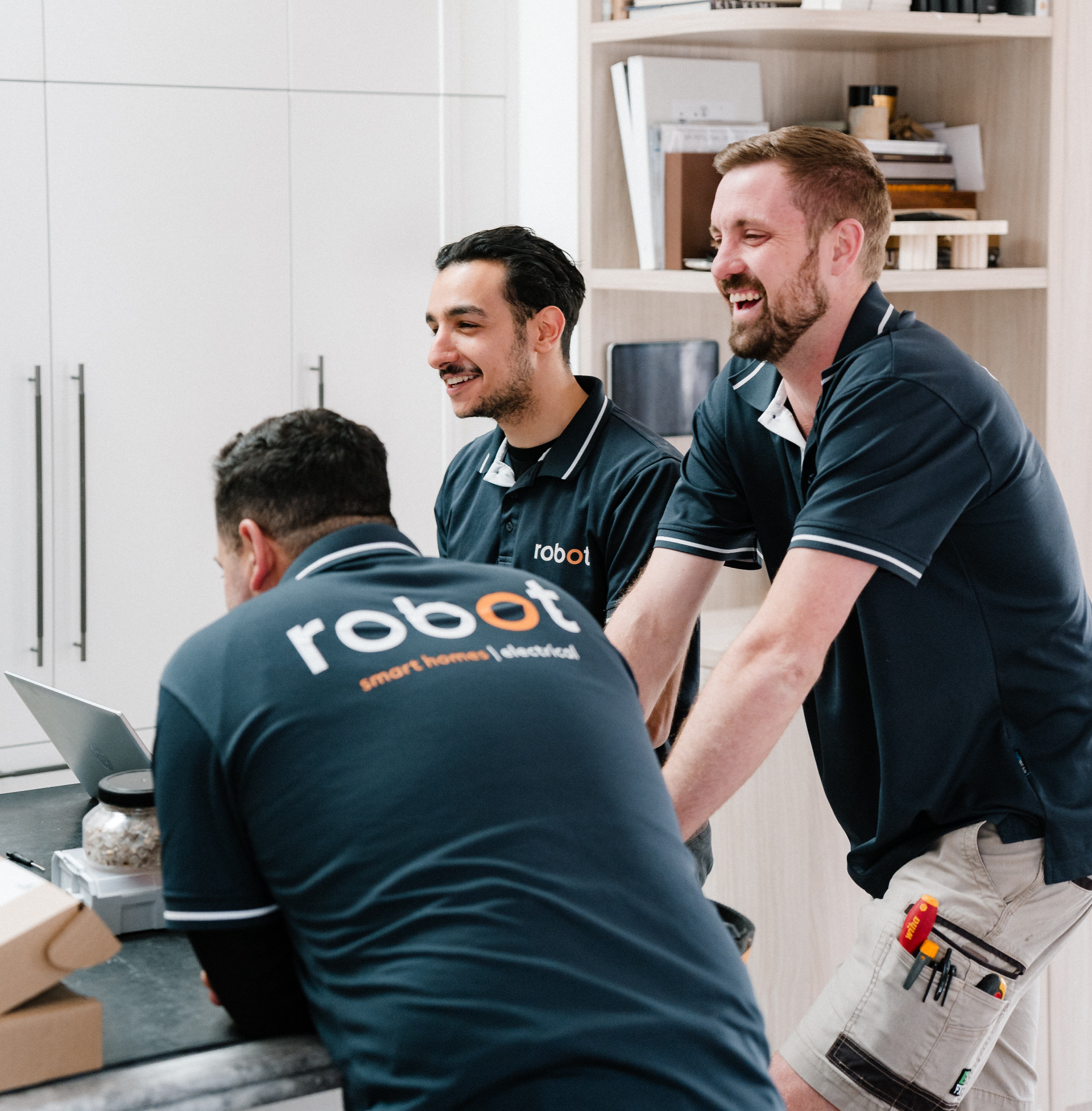 Three men in matching shirts smiling and talking in a modern kitchen.