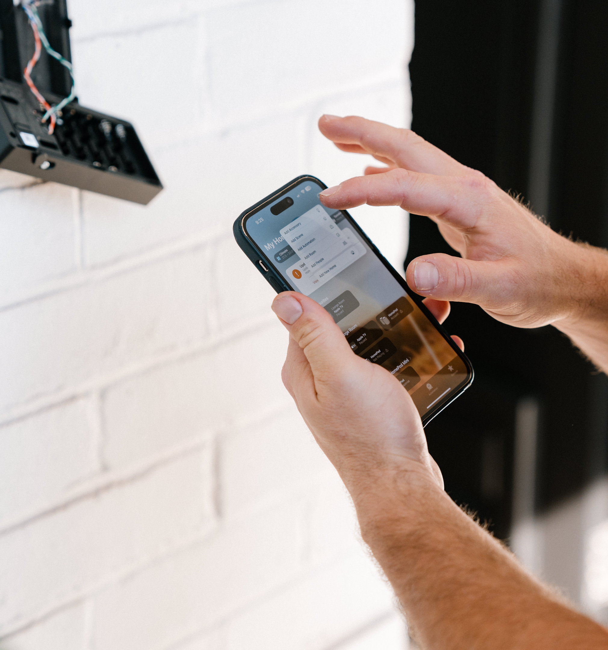 Person using a smartphone near an open electrical box on a white brick wall.