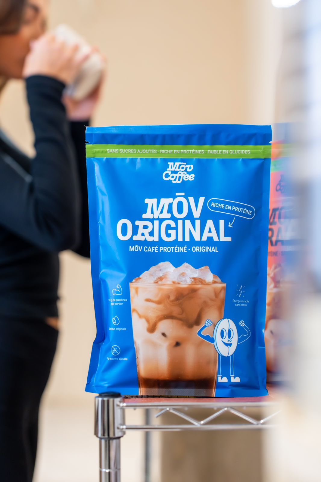 A blue bag of Mōv Original protein coffee mix on a metal shelf with a person drinking behind.