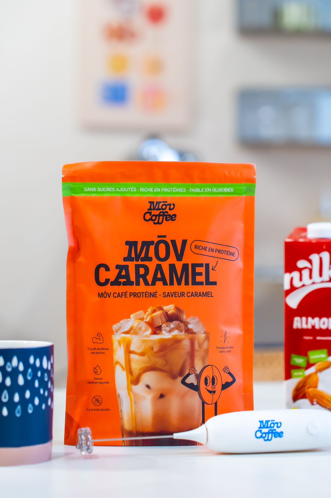 A bag of Mōv Coffee Caramel protein powder sits on a table next to a mug and a frother.