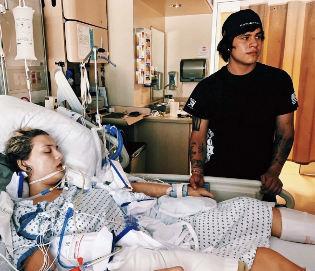 Person in a hospital bed with medical equipment; another person stands beside holding their hand.