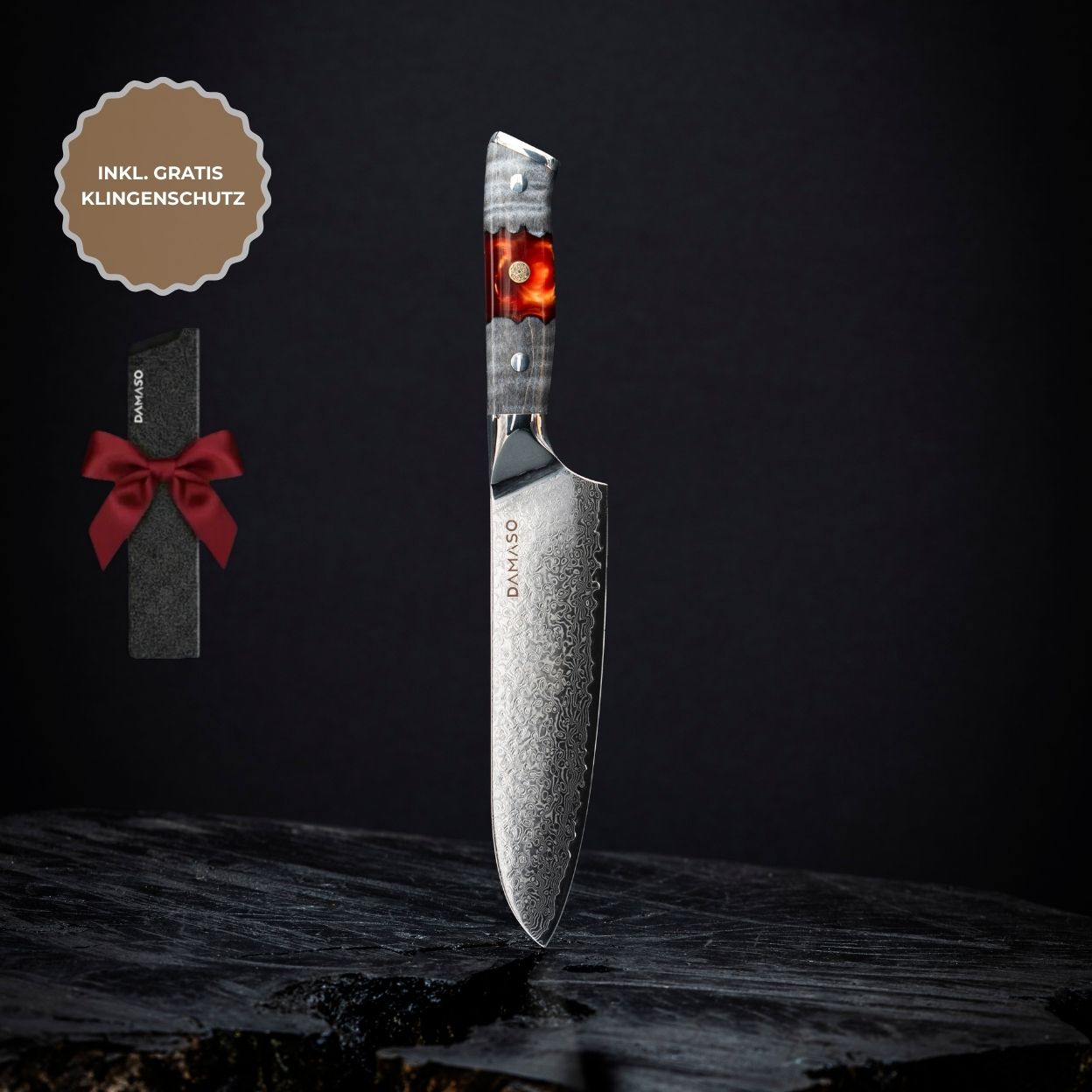 A patterned Damascus knife with a red and grey handle, standing upright on a dark surface beside a gift box.