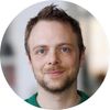 A circular headshot of a man with short brown hair and a light beard, smiling slightly.