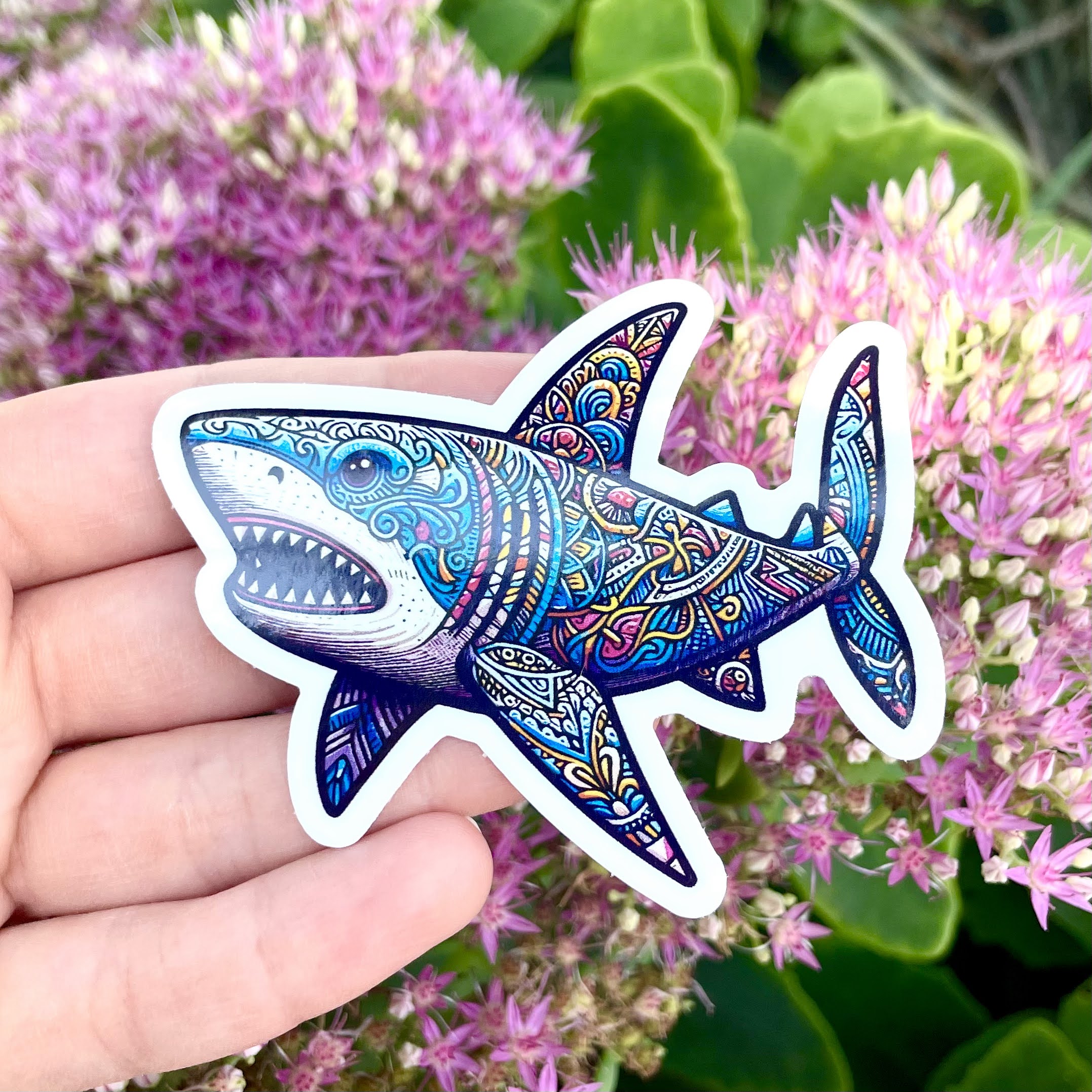 Hand holding a colorful, patterned shark sticker against pink flowers and green leaves background.