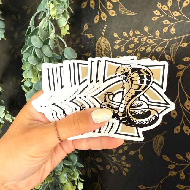 Hand holding stickers featuring a cobra design against a decorative background.