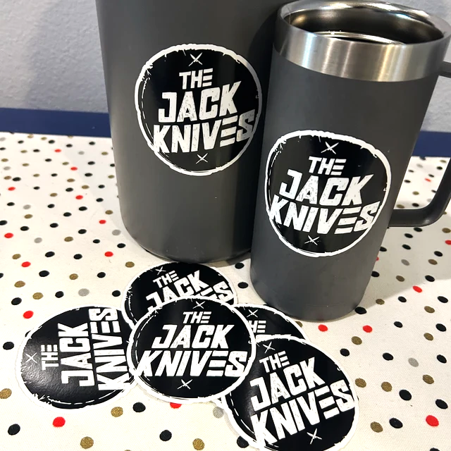 Gray mugs and stickers with 'The Jack Knives' logo on dotted tablecloth.