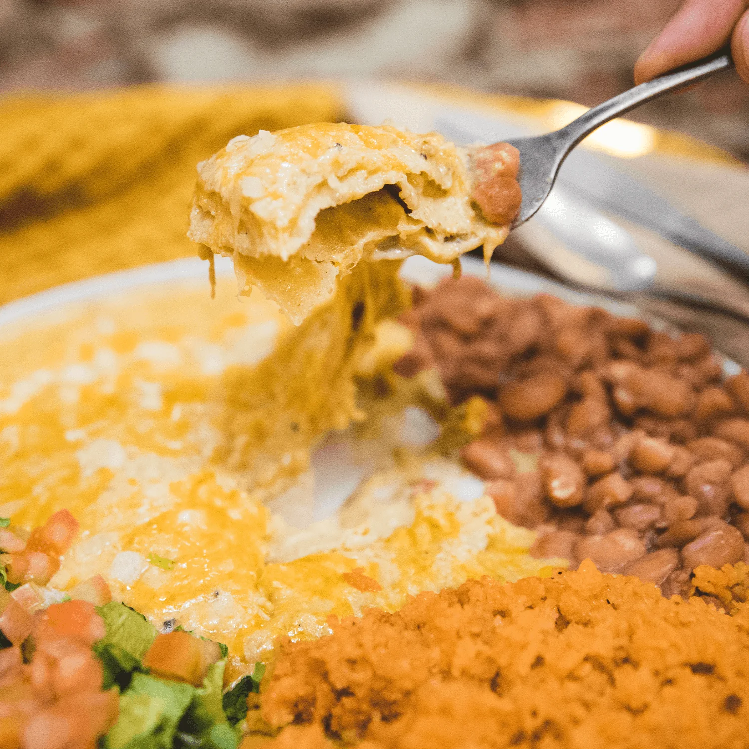 A fork lifts a cheesy bite of food from a plate with rice, beans, and a small salad.