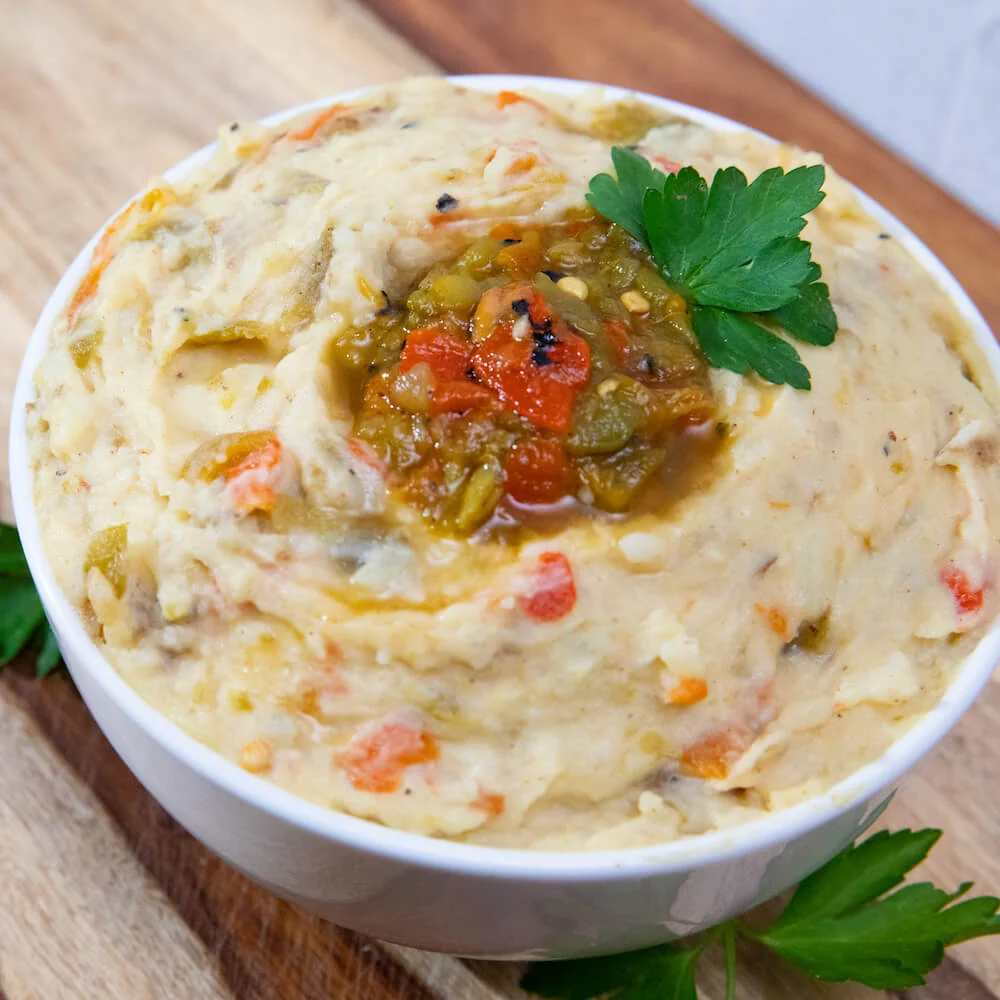 A white bowl of mashed potatoes with chopped vegetables, topped with salsa and a parsley leaf on a wood board.