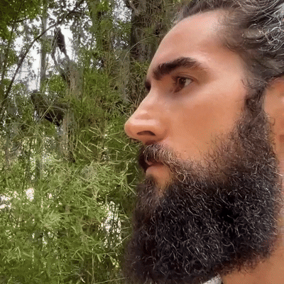 Profile of a bearded man outdoors with greenery in the background.
