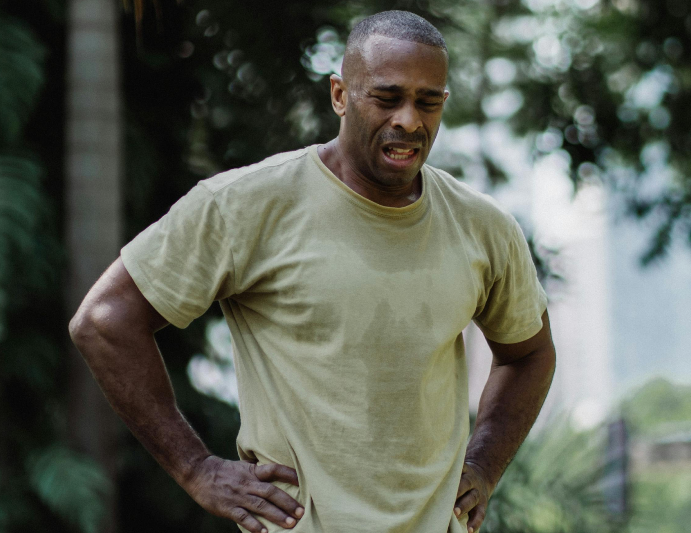 Man in a beige shirt appears tired, standing outdoors with hands on hips.